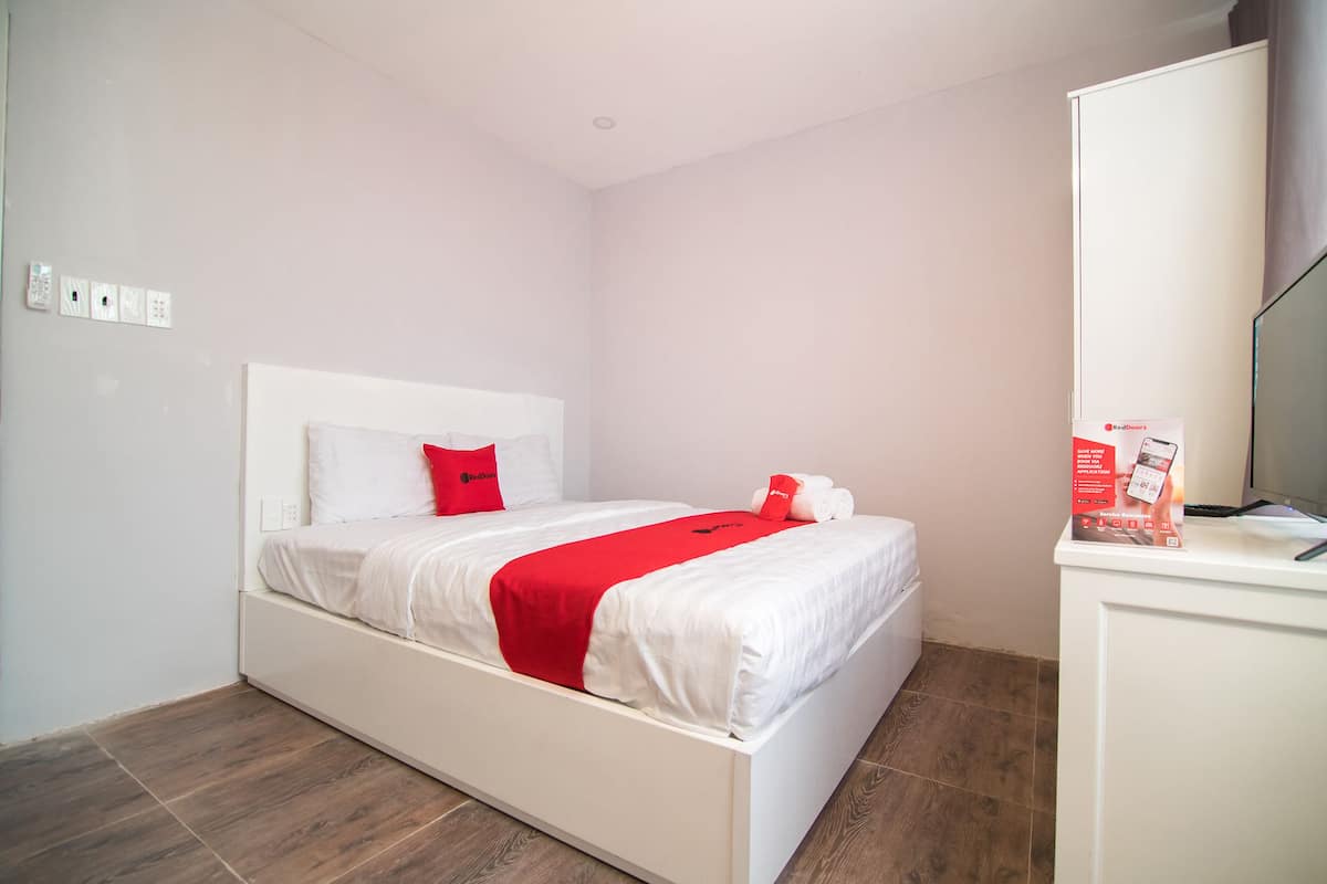 A neatly made bed with white bedding and a red accent pillow, a small bedside table with a red box, and a white wardrobe.