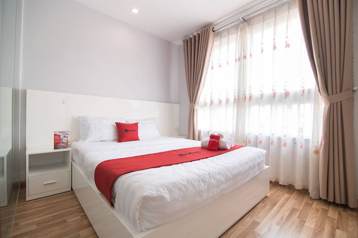 A neatly made bed with a red blanket and pillows, a bedside table with a RedBook magazine, and a window with floral curtains.