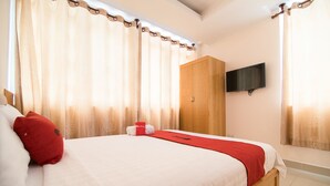 Un lit bien fait, agrémenté d’un coussin rouge, une armoire en bois et un téléviseur à écran plat fixé au mur.