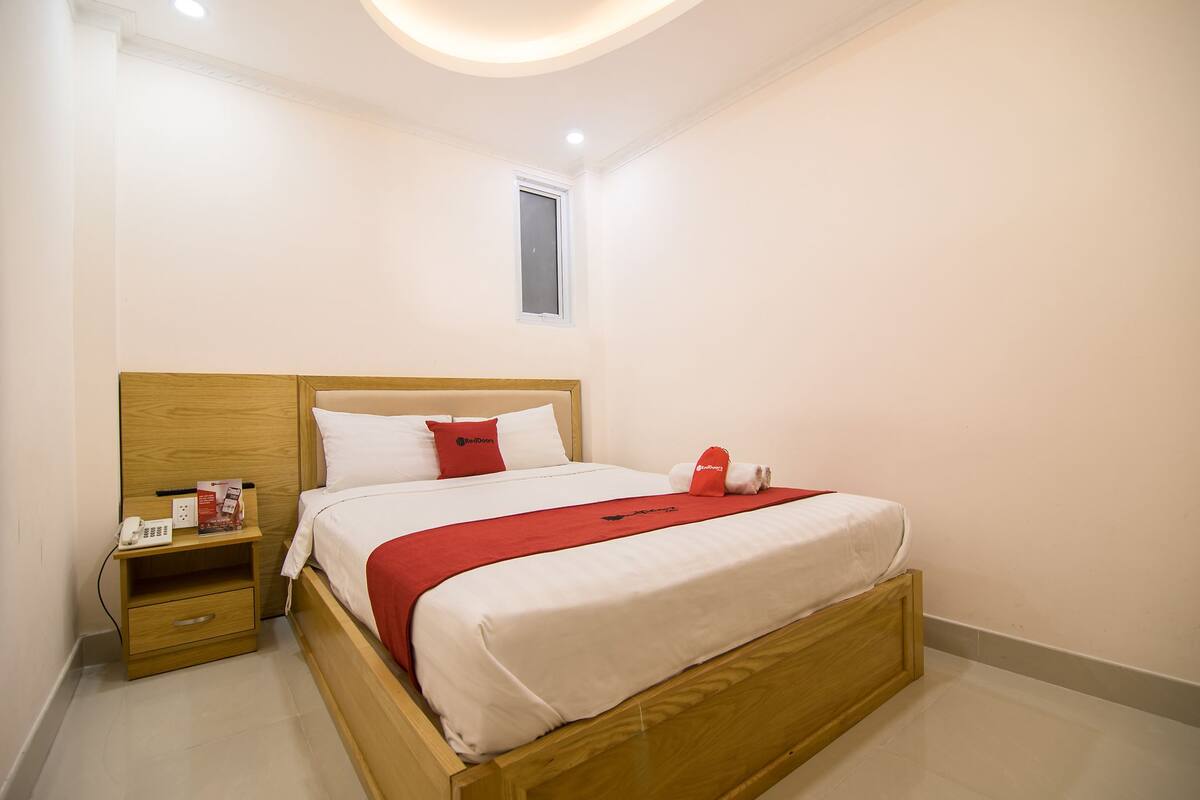 Una cama bien tendida con cabecera de madera, una mesita de noche con un teléfono y folletos, y una ventana pequeña sobre la cama.