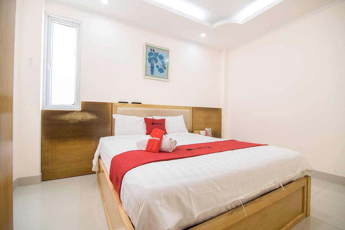 Una cama bien tendida con sábanas blancas y una manta roja, un cabecero de madera y una mesita de noche con una lámpara.