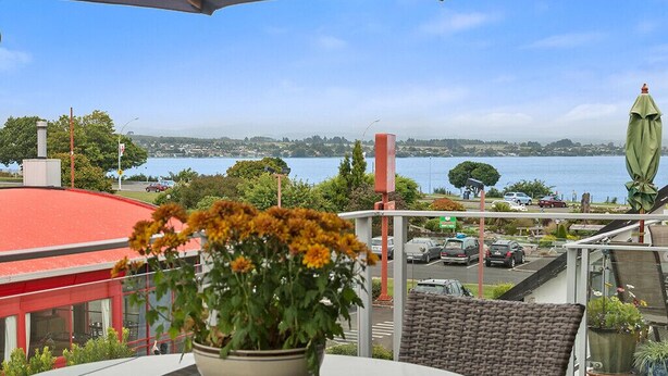 A terrace with a view of a body of water, a parking area, and a building with a red roof.