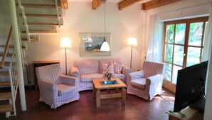 A cozy living room with a sofa set, two armchairs, a coffee table, and a TV. There is a staircase on the left and a window on the right.