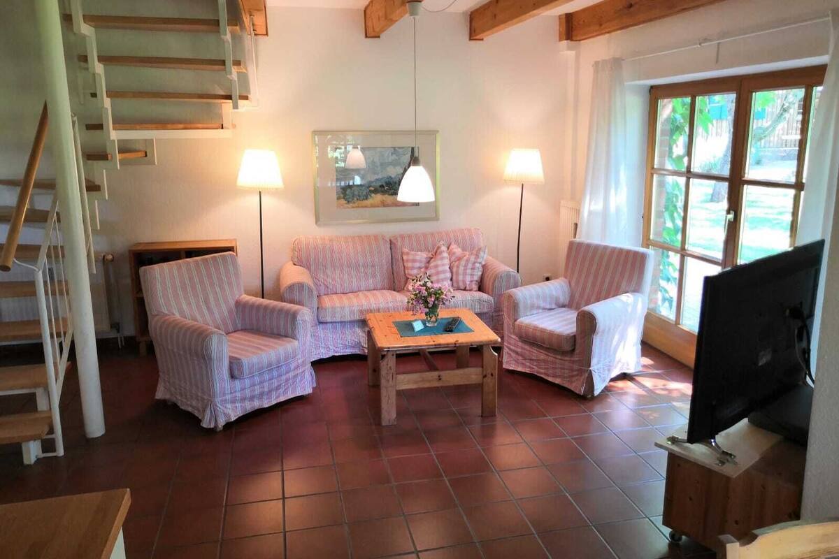 Een gezellige woonkamer met een zithoek, twee fauteuils, een salontafel en een televisie. Links een trap en rechts een raam.