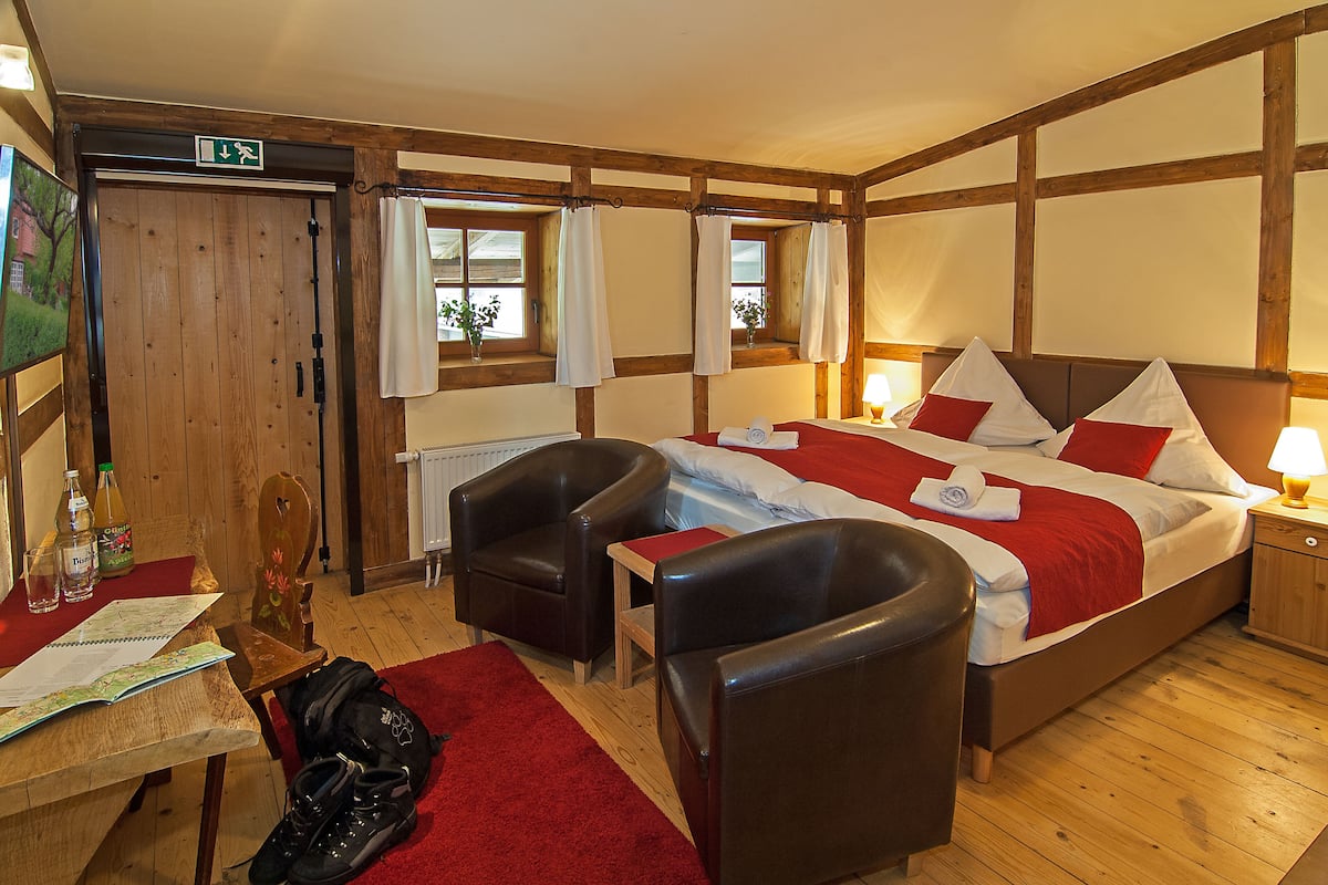Een kamer met houten lambrisering, een bed, twee stoelen, een tafel met een kaart en een rode vloerkleed.