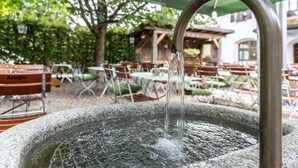 An outdoor seating area with tables and chairs, a water feature, and a green umbrella with a logo. - Hotel Alter Hof (Vaterstetten)