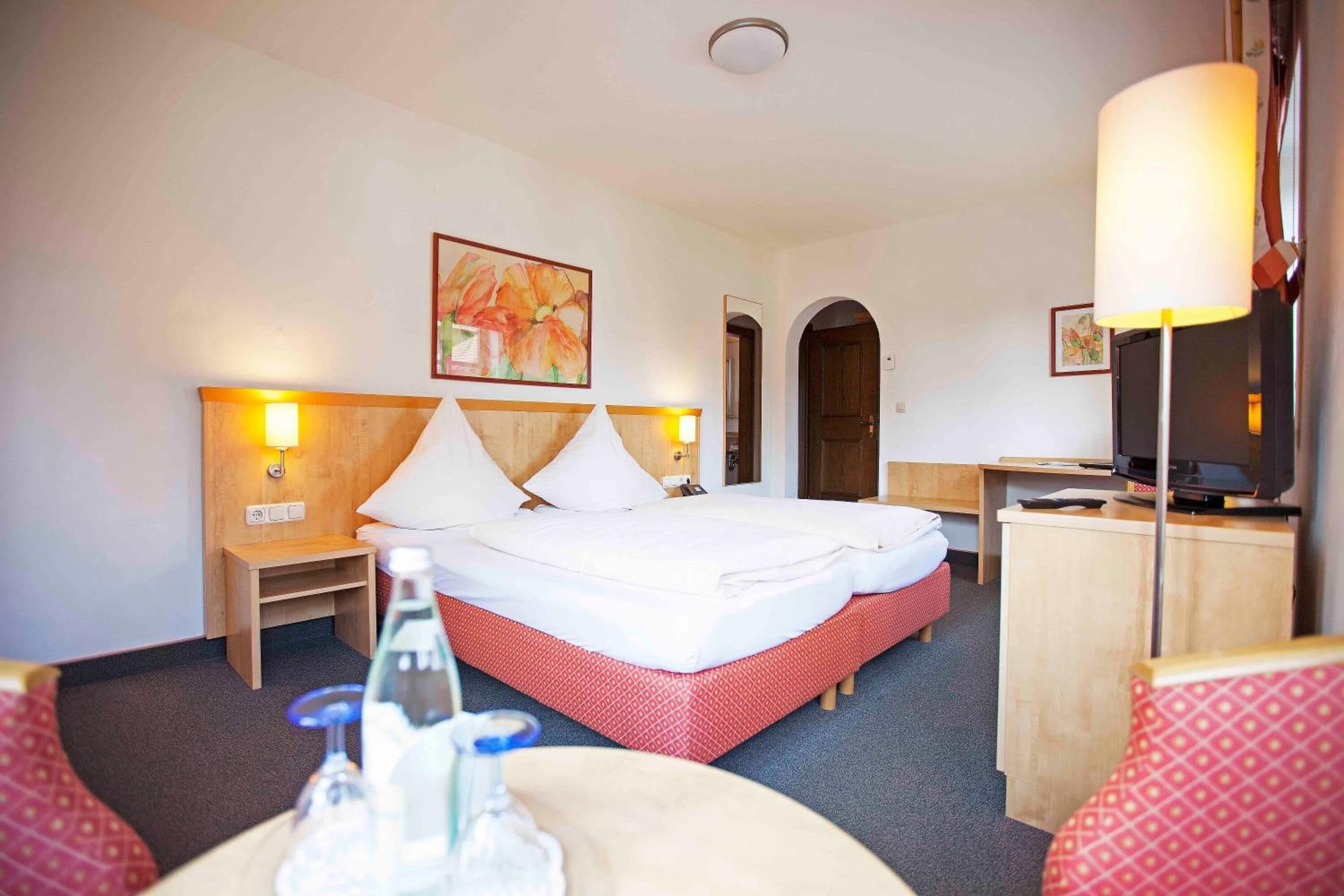 A hotel room with a bed, bedside tables, a desk, a television, and a painting on the wall.