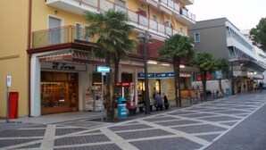 A multi-story building with balconies, a storefront with a sign, and a pedestrian area with palm trees and benches. - App'artamento Soyus n.10 (Jesolo)