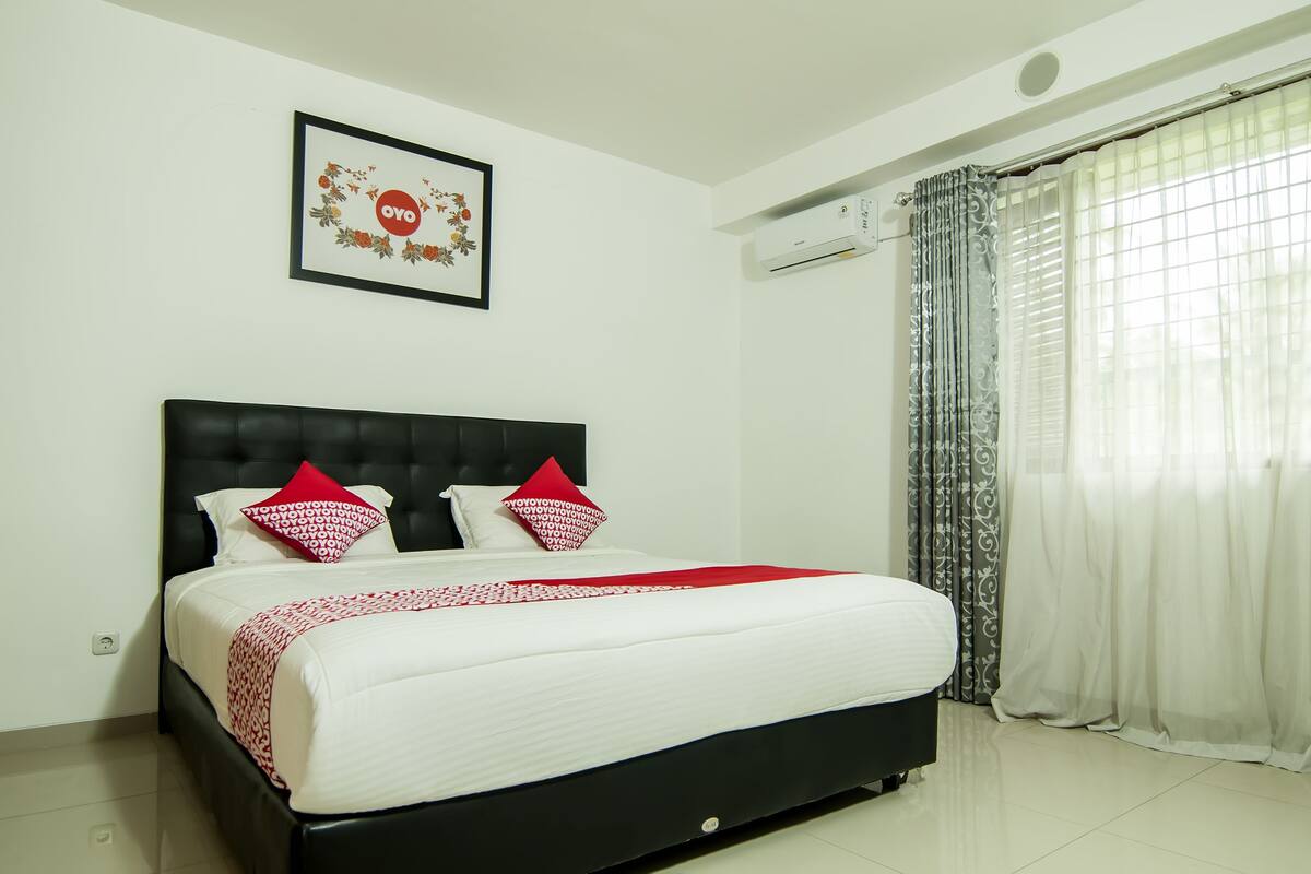 A neatly made bed with a black headboard, white bedding, and red decorative pillows. There is a framed picture on the wall above the bed, and an air conditioning unit mounted on the wall.