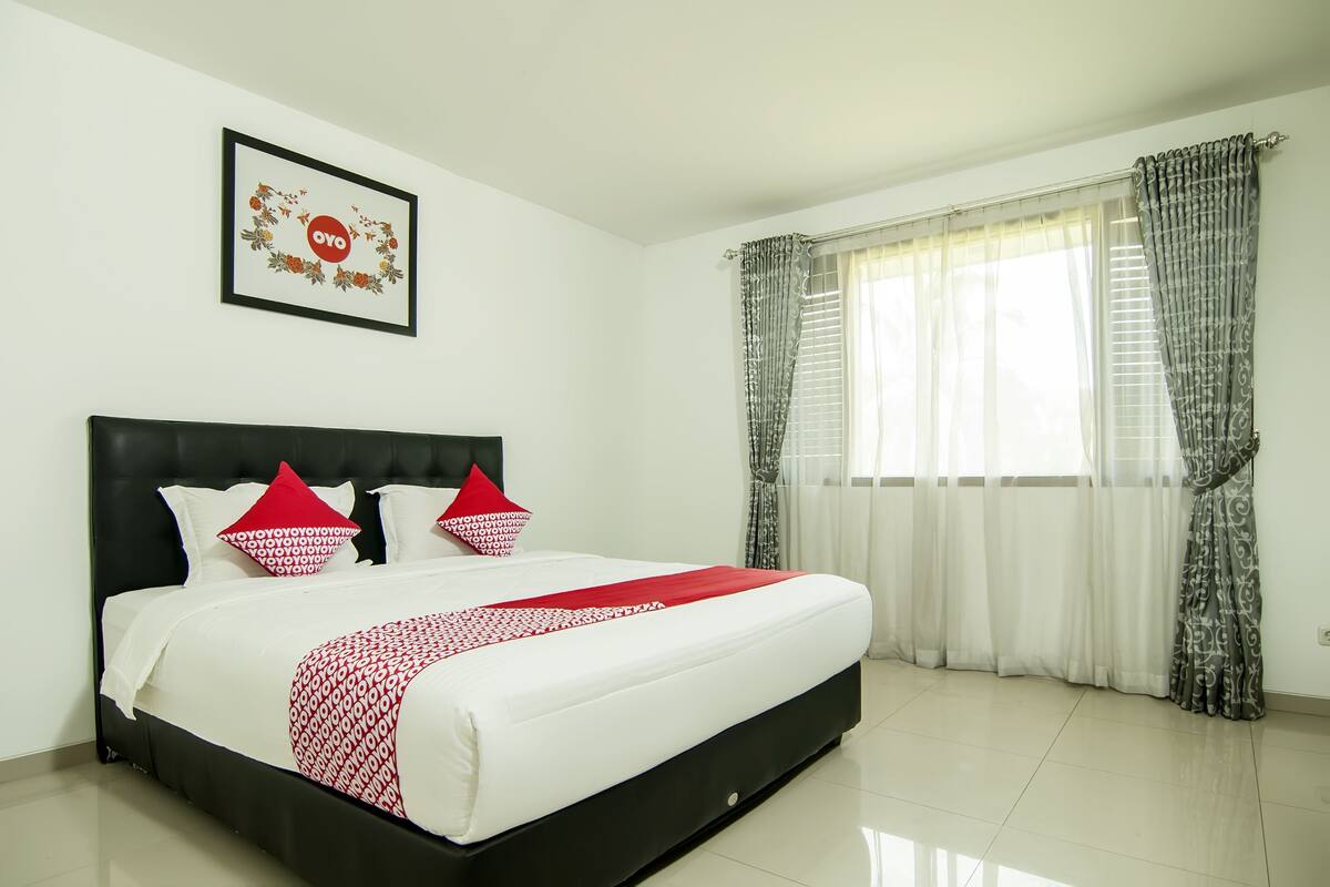 A neatly made bed with a black headboard, white bedding, and red pillows. There is a framed picture on the wall above the bed.
