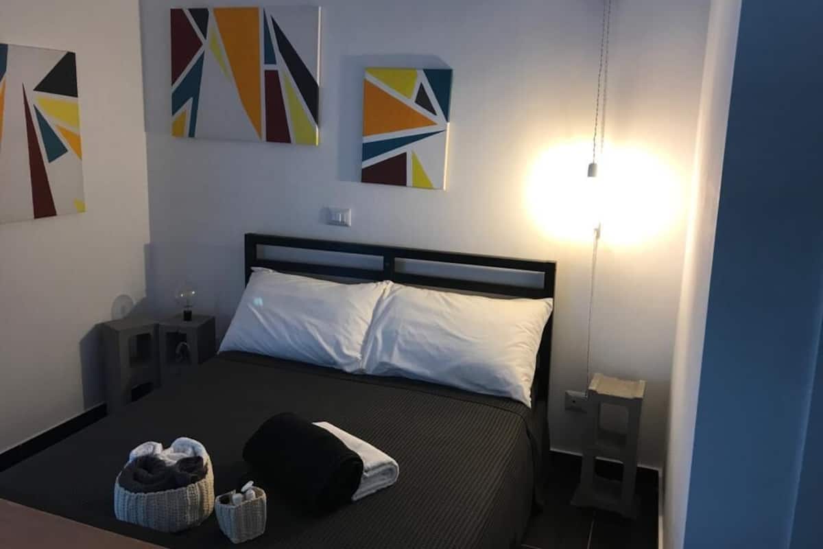 A neatly made bed with white pillows and a dark bedspread, a bedside table with a lamp, and abstract wall art.