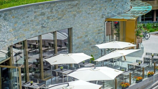 Een terras met glazen tafels en witte parasols, omgeven door stenen muren en begroeiing.