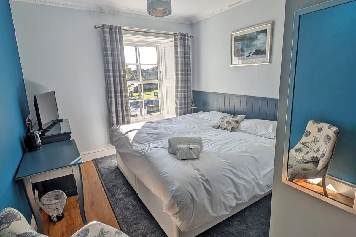 A bedroom with a bed, a desk, a chair, a window with curtains, and a framed picture on the wall.