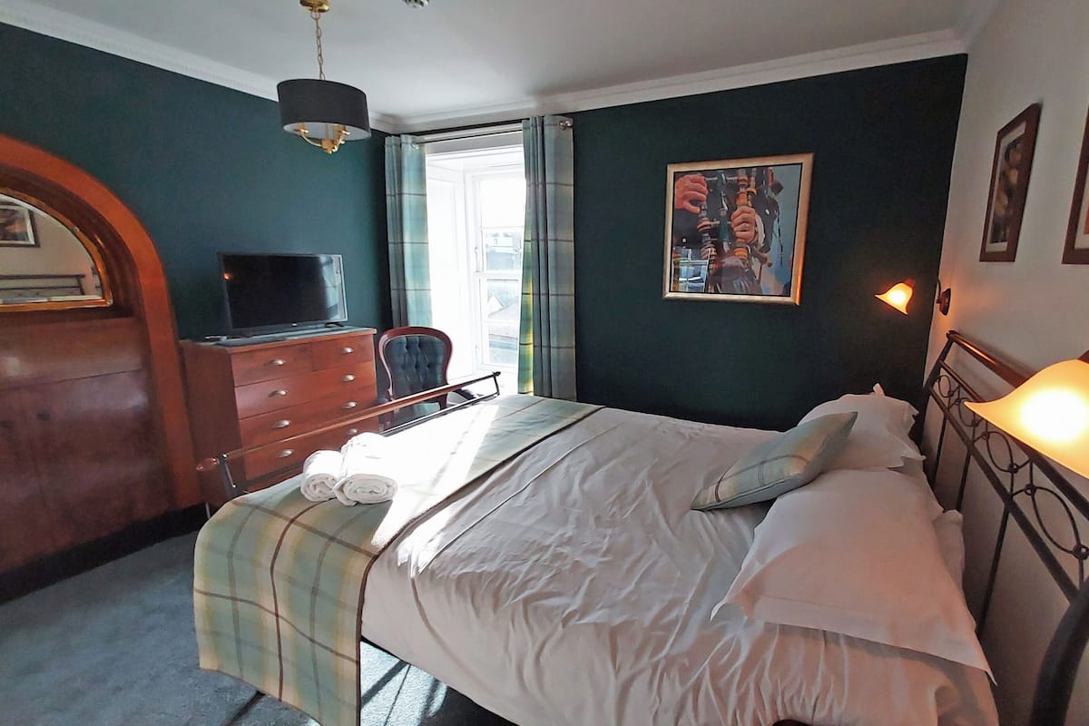 A bedroom with a bed, a desk, a chair, a television, and a painting on the wall.