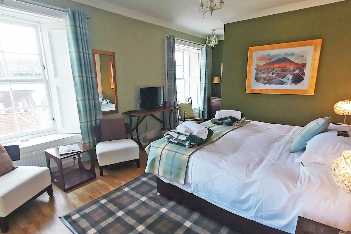 A hotel room with a bed, a desk, a chair, a television, and a painting on the wall.