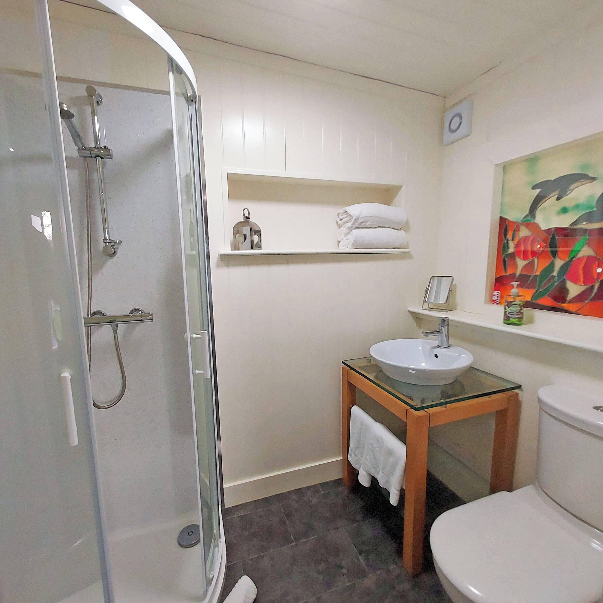 A bathroom with a glass shower enclosure, a glass-top sink, a toilet, and a shelf with towels.