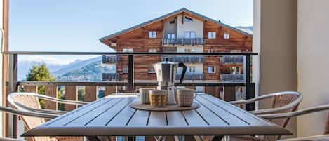 A balcony with a wooden table and wicker chairs, overlooking a mountain and a building with balconies.