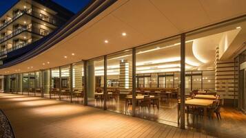 A modern hotel exterior with a glass facade, outdoor seating area, and a curved roof structure.
