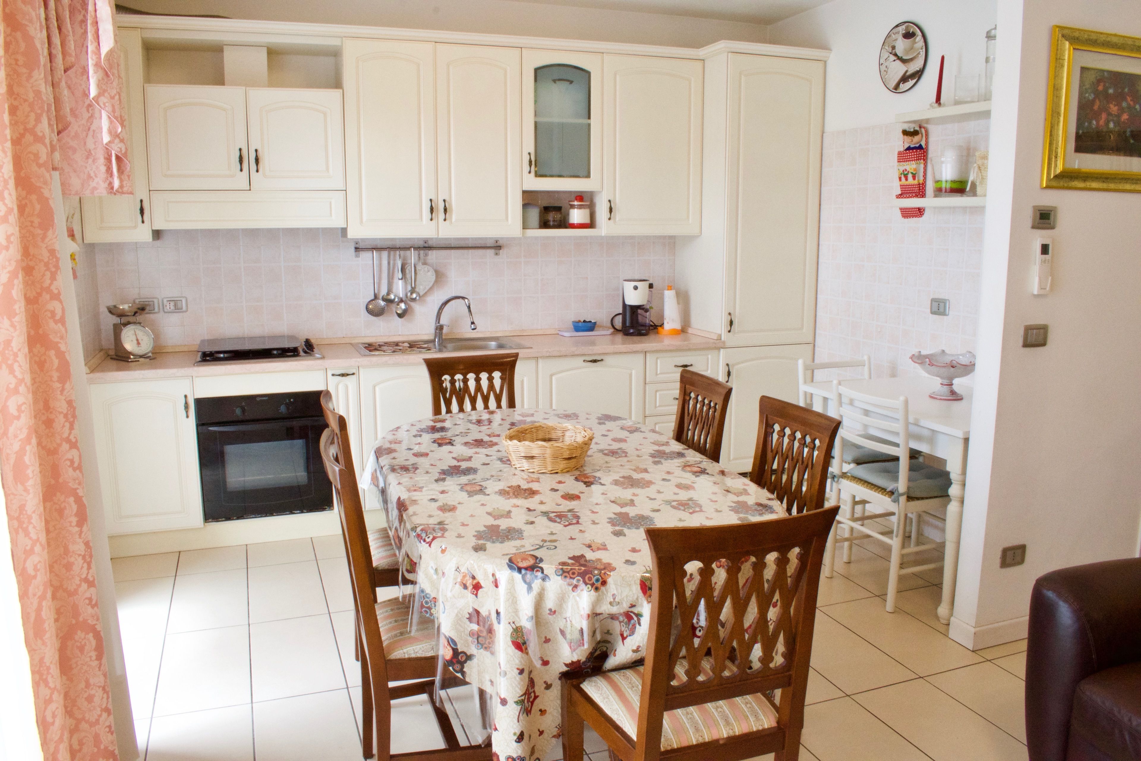 A kitchen with white cabinets, a dining table with floral tablecloth, wooden chairs, and a coffee maker.