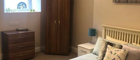 A bedroom with a bed, a wardrobe, a nightstand, a lamp, and a window with a view of a car.