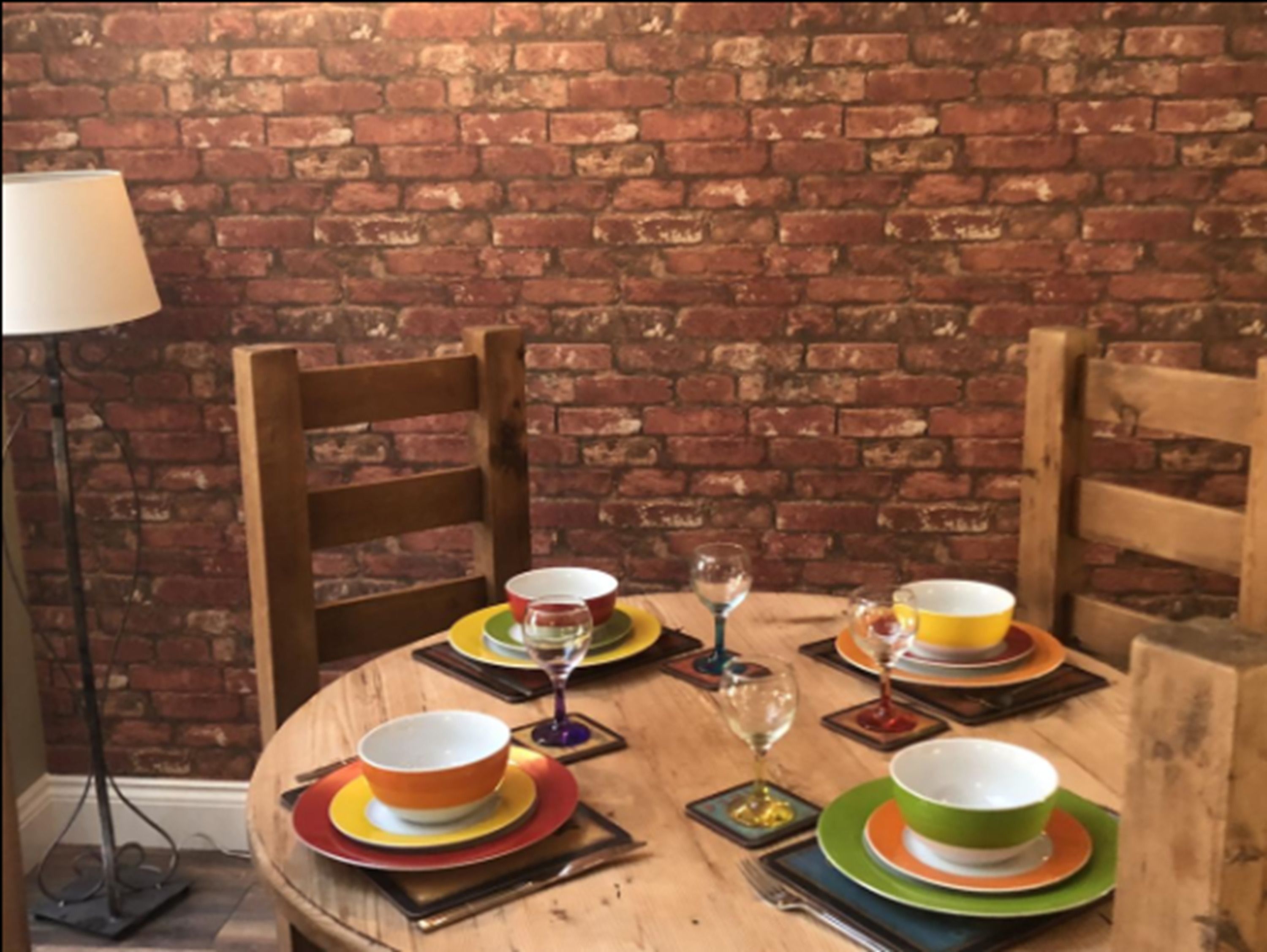 Een ronde eettafel met kleurrijke borden en glazen, voor een bakstenen muur.
