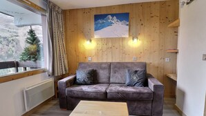 Una habitación acogedora con un revestimiento de madera, un sofá gris con cojines decorativos, una mesita de madera y una ventana con vistas a una montaña nevada.