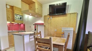 Cocina compacta con armarios de madera, microondas, televisor montado en la pared y comedor con sillas y mesa de madera.