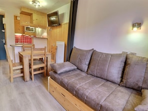 A compact living space with a sofa, wooden dining table, chairs, a TV mounted on the wall, and a kitchenette area.