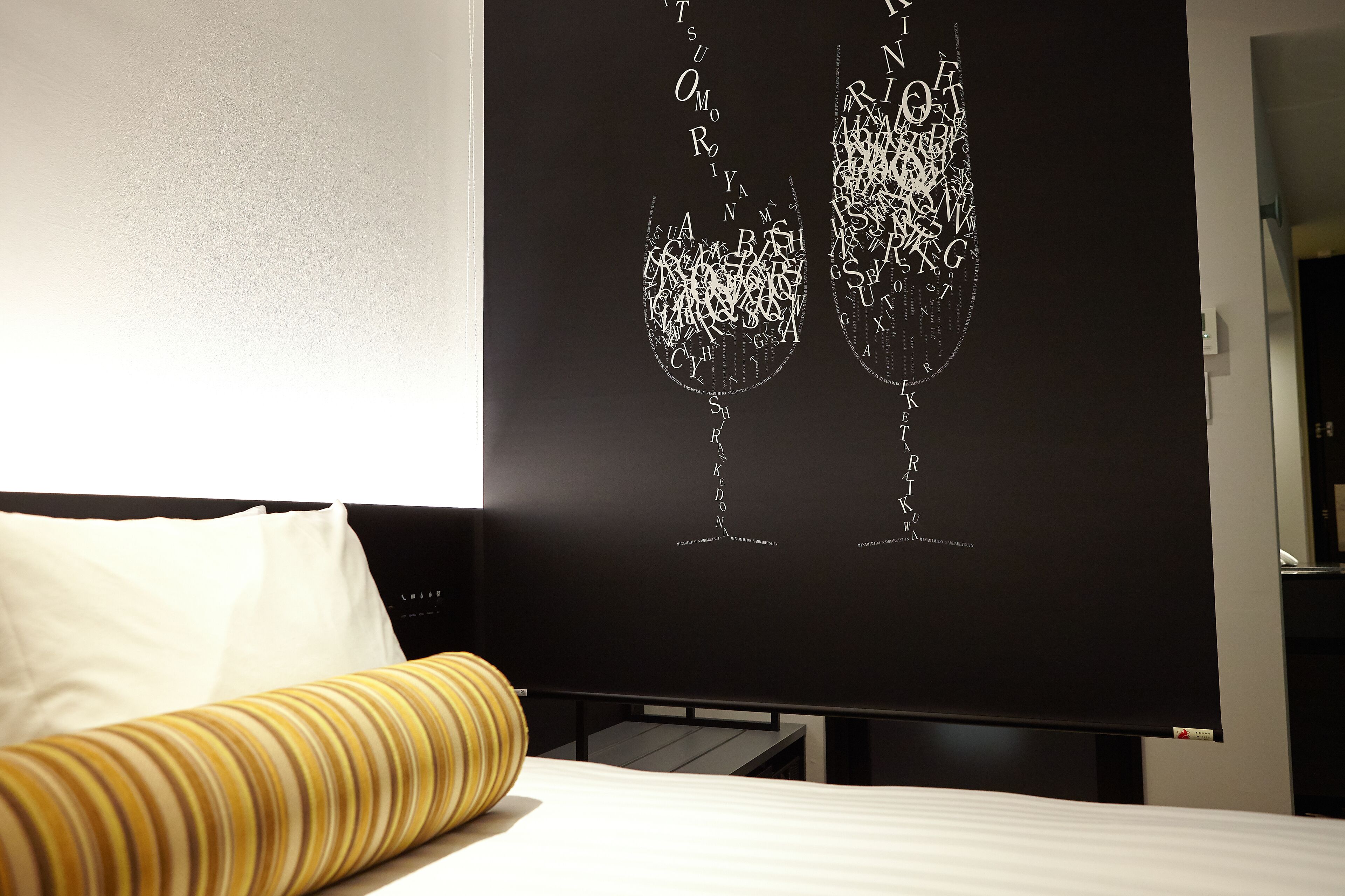 A hotel room with a bed, a large black artwork of two wine glasses, and a yellow striped pillow.