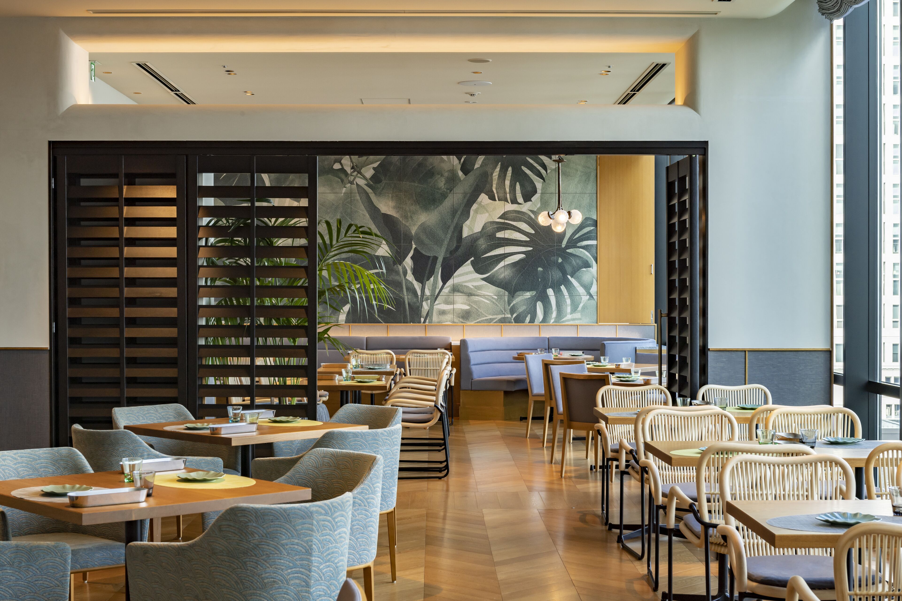 A modern restaurant interior with wooden tables and chairs, a large tropical mural, and a seating area with cushioned benches.