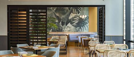 A modern restaurant interior with wooden tables and chairs, a large tropical mural, and a seating area with cushioned benches.