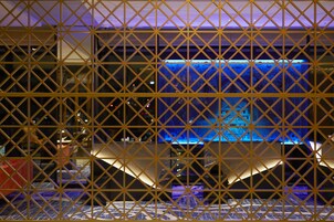 A room with a geometric patterned partition, a bed with a blue patterned bedspread, and a large window with a view of a building.