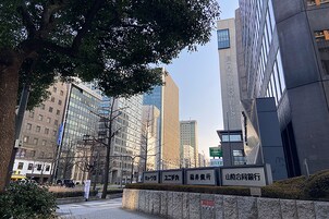 A modern urban street with tall buildings, a tree-lined sidewalk, and a stone wall with signage.