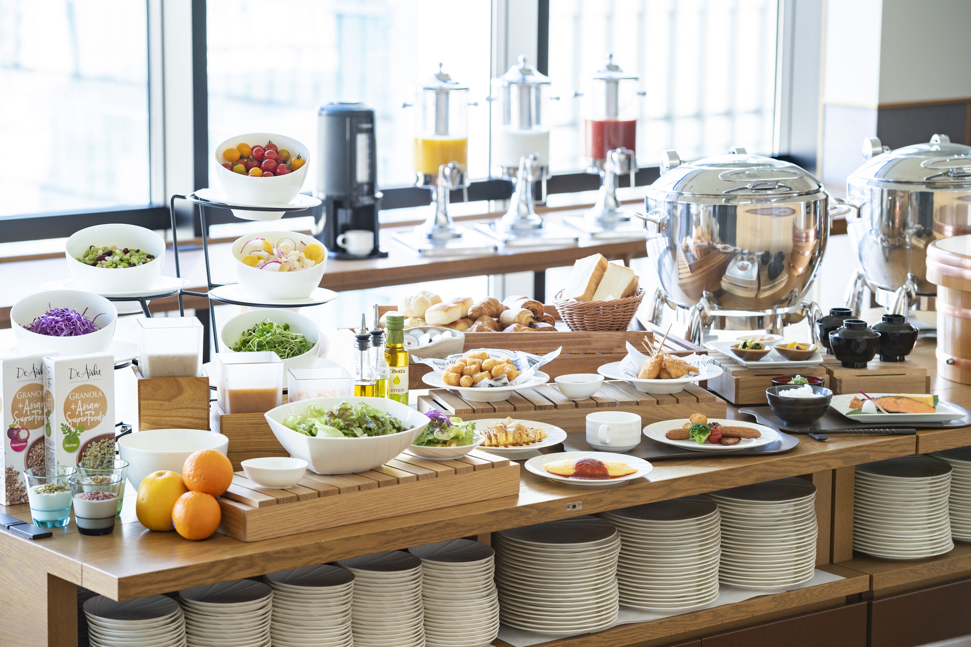 A buffet setup with various food items, including fruits, salads, and bread, arranged on a wooden table with stacked plates and bowls.