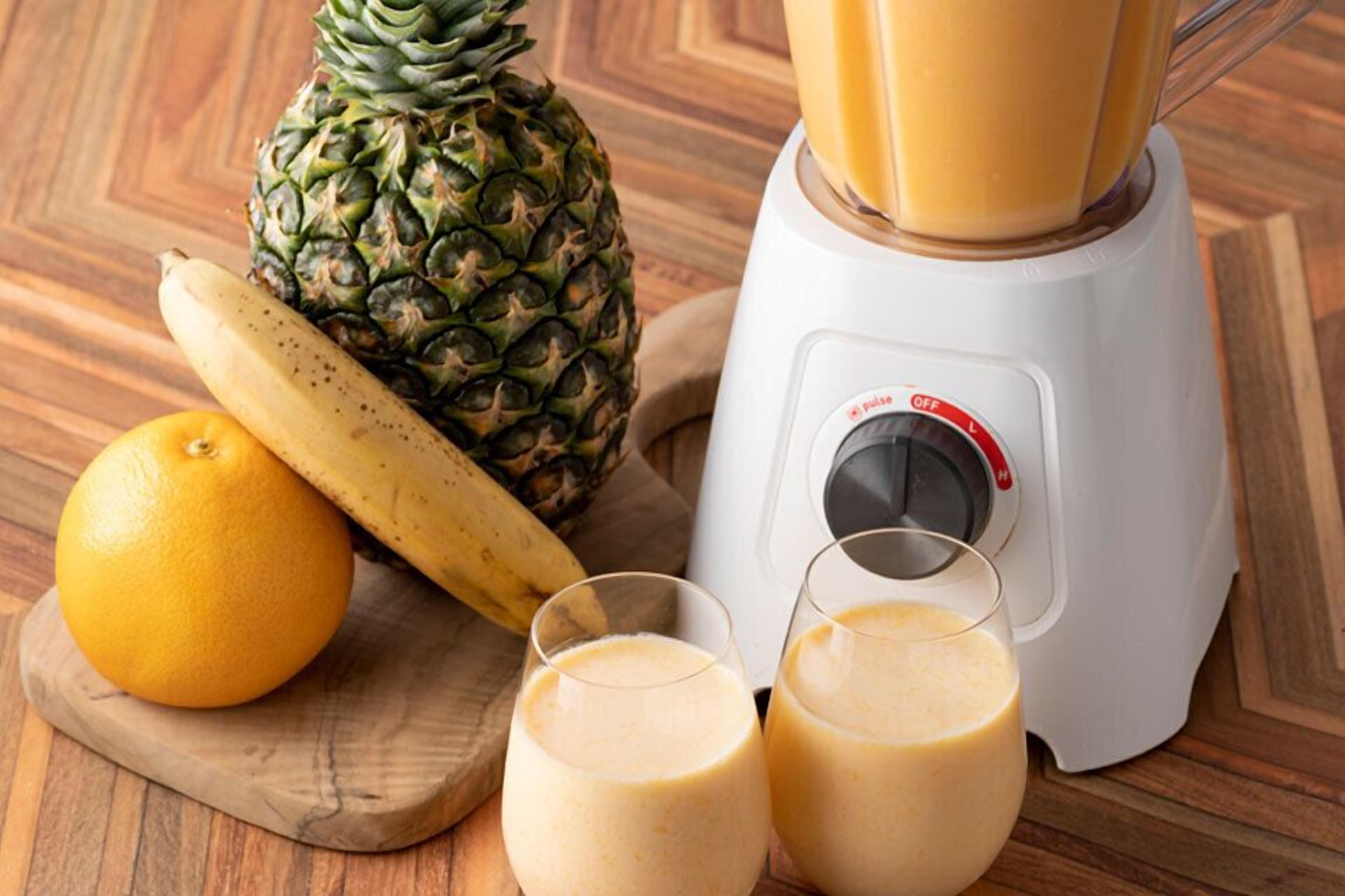 Una licuadora con dos vasos de batido, una piña y un plátano sobre una superficie de madera.