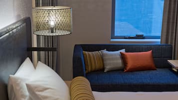 Una habitación de hotel con una cama, un sofá azul con cojines, una lámpara y una ventana con vistas.