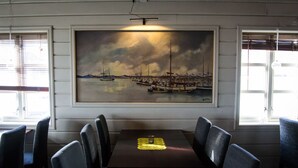 A dining area with a table and chairs, a painting of a harbor scene, and windows with blinds.