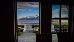 A view from inside a room with a large window overlooking a wooden deck with benches and a railing, leading to a body of water and snow-capped mountains in the distance.