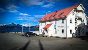 A white building with a red roof, labeled "Brygga Hotel Lodingen," located near a body of water with mountains in the background.
