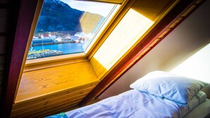 A small, single-bed room with a window overlooking a body of water and a mountainous landscape.