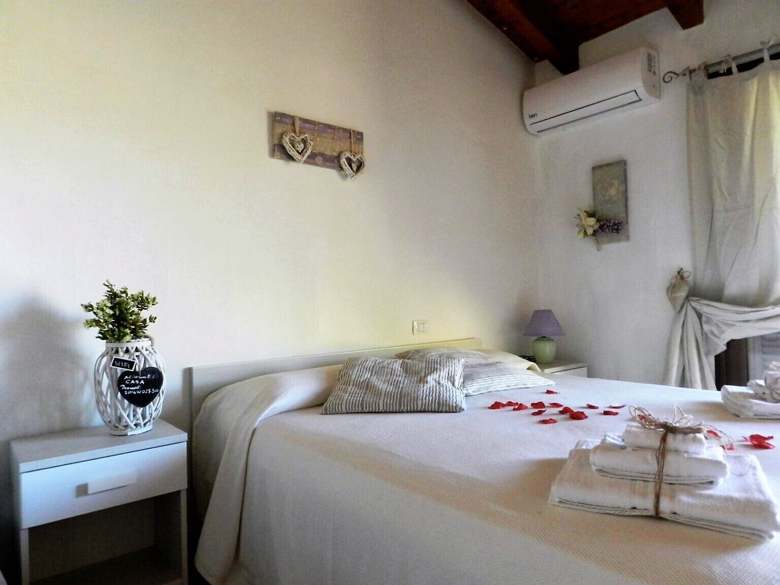 Una cama bien hecha con un almohadón a rayas, un pequeño florero con una planta y una decoración con forma de corazón en la pared.