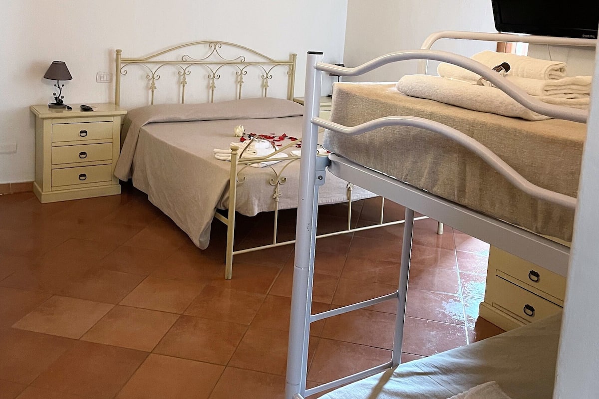 A bedroom with a bunk bed, a bedside table with a lamp, a bed with a white comforter, and an air conditioning unit on the wall.
