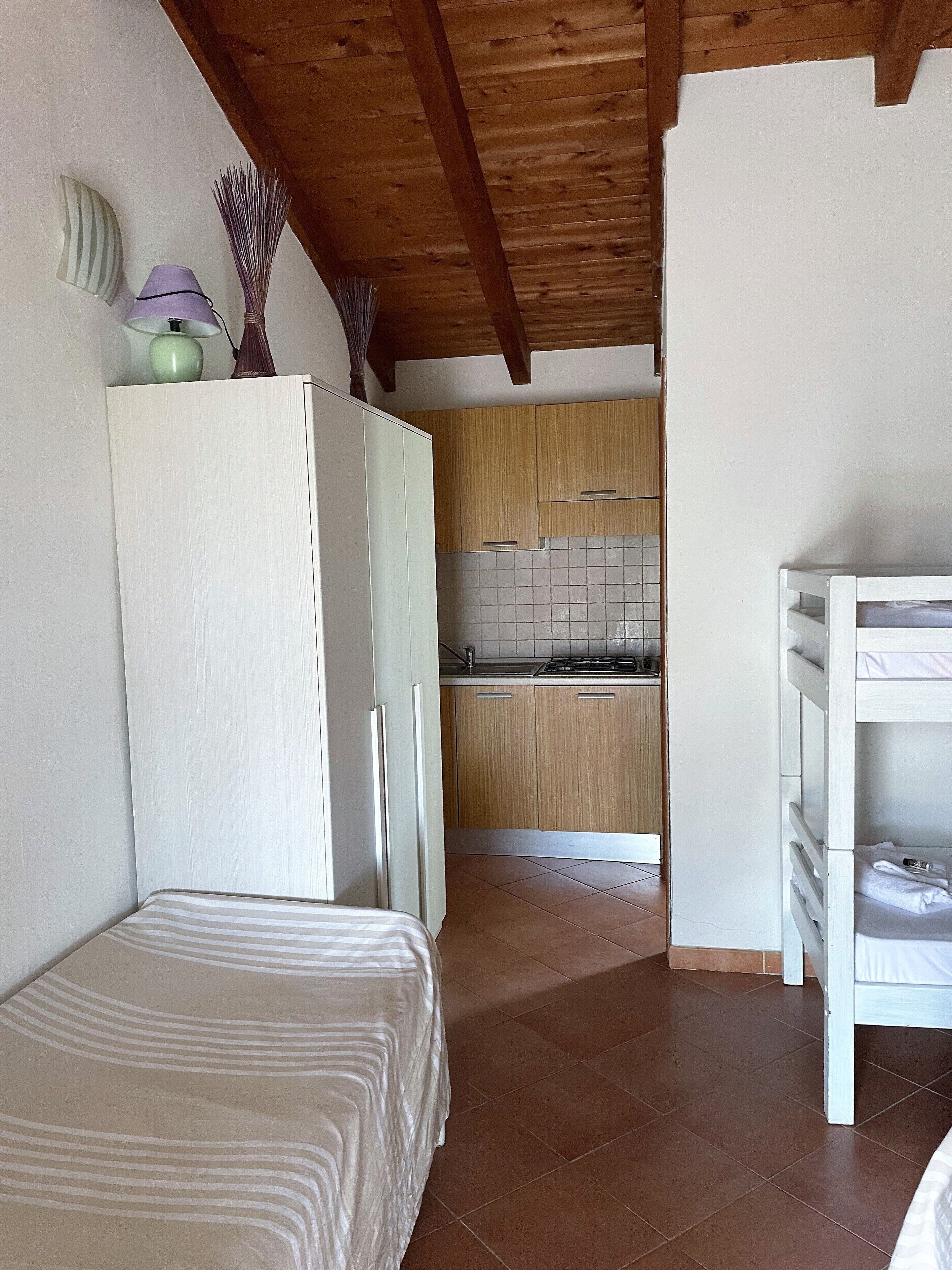 A bedroom with a bed, a white wardrobe, and a small kitchenette with a tiled backsplash and wooden cabinets.