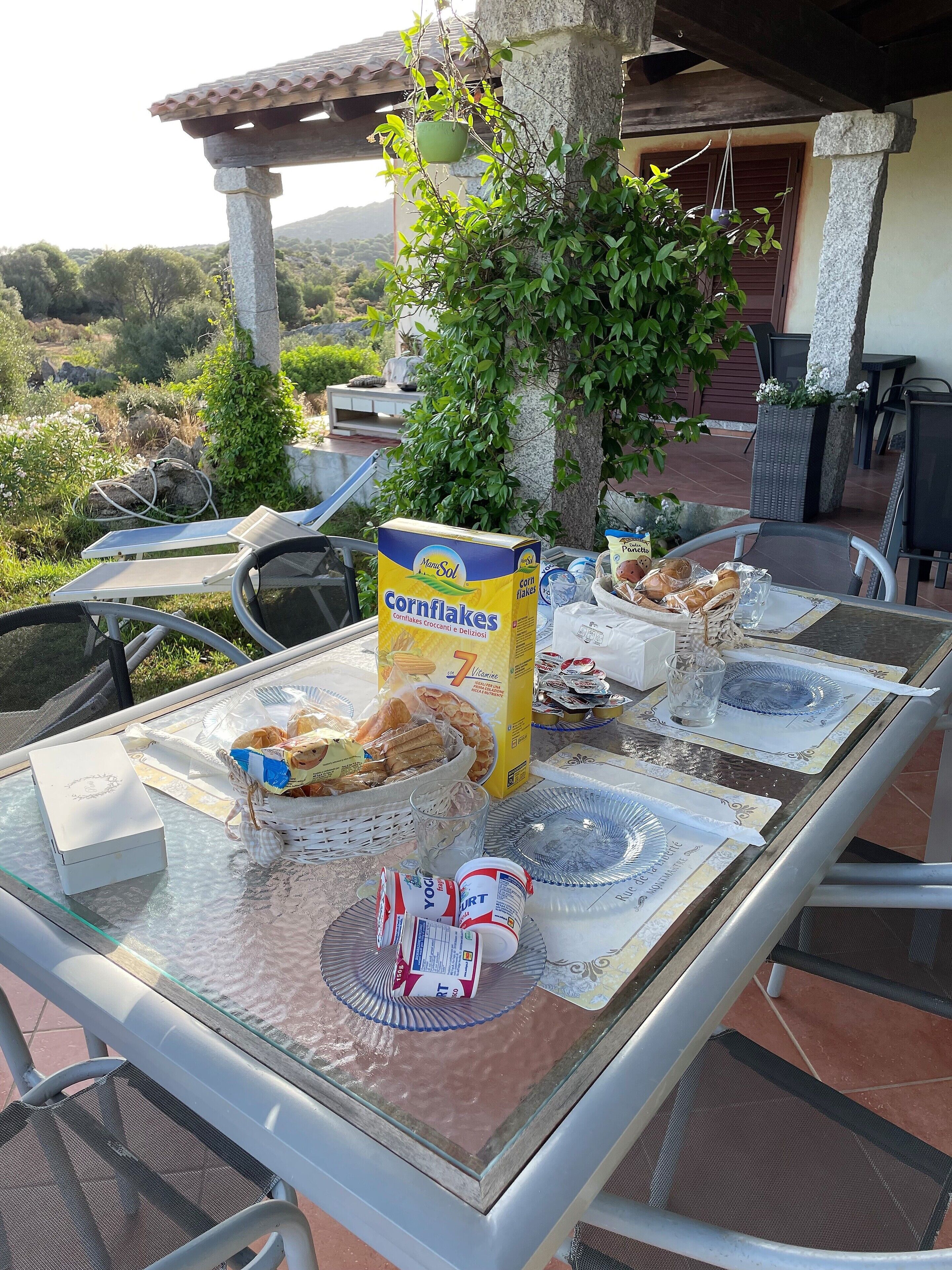 A table set outdoors with a box of cornflakes, plates, glasses, and a basket of bread.