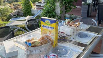 A table set outdoors with a box of cornflakes, plates, glasses, and a basket of bread.