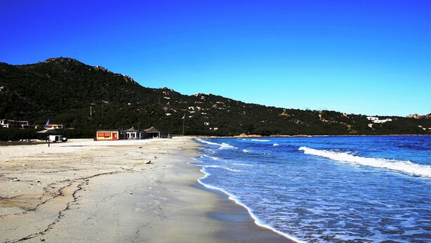 A beach with a clear sky, waves, and buildings in the background. - B&B Su Donnigheddu (Golfo Aranci)