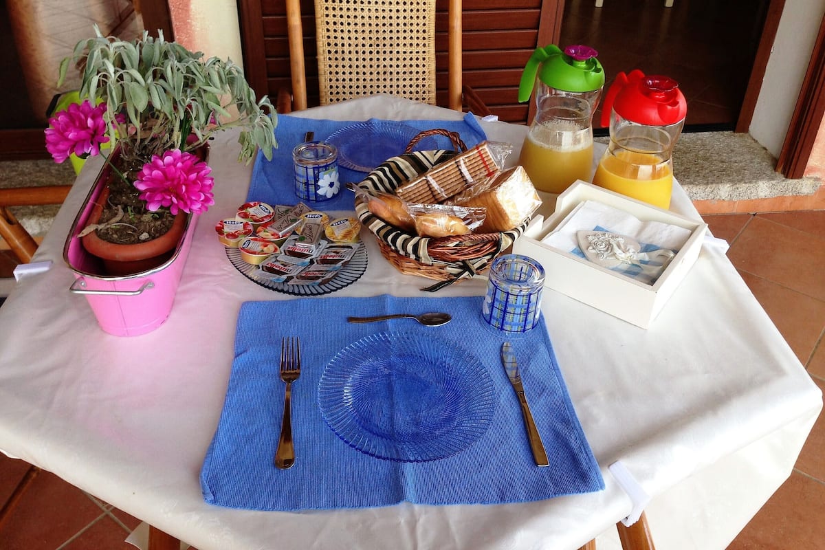 A breakfast setup with a table set for two, including plates, cutlery, a blue placemat, a potted plant, and a basket of bread.