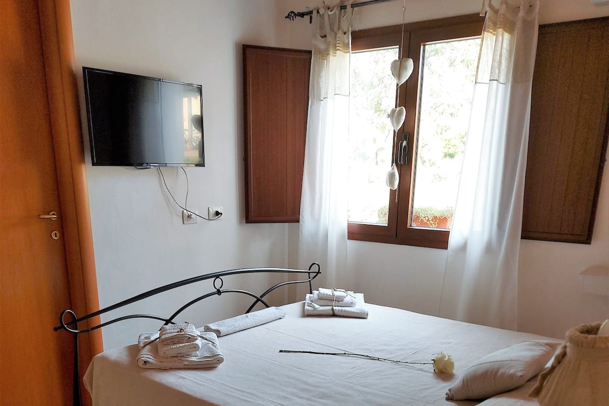 A neatly made bed with towels, a bedside table, a TV mounted on the wall, and a window with curtains.