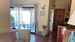 A cozy living room with a sofa, two small tables, a potted plant, and a kitchen area with a refrigerator and a lamp. - B&B Su Donnigheddu (Golfo Aranci)