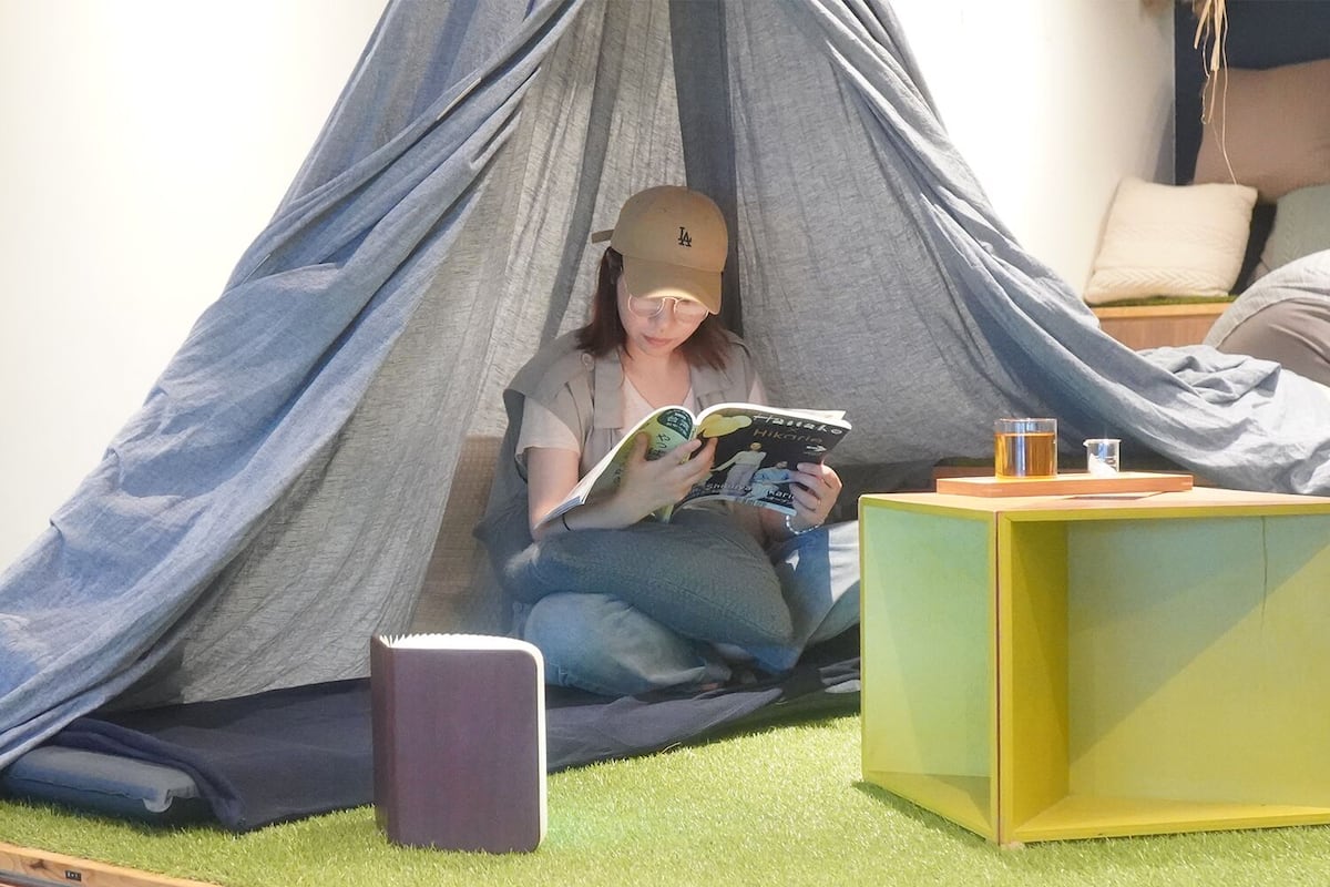 A person reading a book in a tent-like structure with a small table and a trash bin nearby.